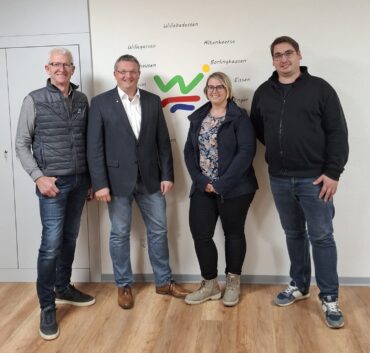 Drei Männer und eine Frau stehen vor einer weißen Wand in einem Raum. Auf der Wand ist das Logo der Stadt Willebadessen und die Namen der Ortsteile.