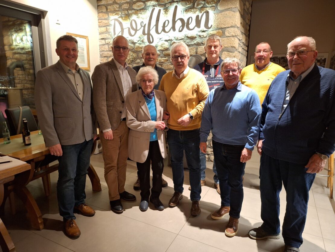 8 Männer und eine Frau stehen vor einer Bruchsteinwand mit einem beleuchteten Schriftzug Dorfleben.