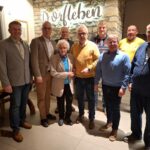 8 Männer und eine Frau stehen vor einer Bruchsteinwand mit einem beleuchteten Schriftzug Dorfleben.