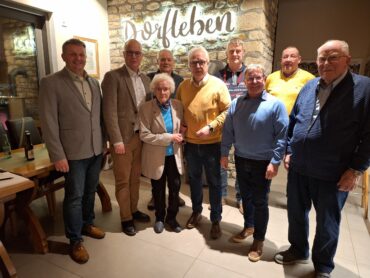 8 Männer und eine Frau stehen vor einer Bruchsteinwand mit einem beleuchteten Schriftzug Dorfleben.